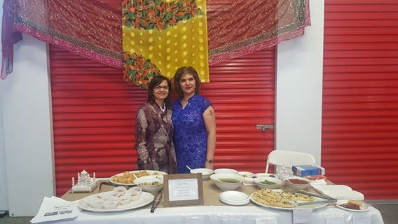 Flavours of Thorncliffe, two women standing behind a white rectangular table with several plates of food on top, two red storage units in th eback